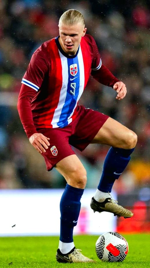 A male soccer player in a red and blue striped jersey controls a ball on the field during an IPTV Latino-broadcasted match. He has blond hair tied back, maroon shorts, and blue socks. The background is blurred with stadium lights.