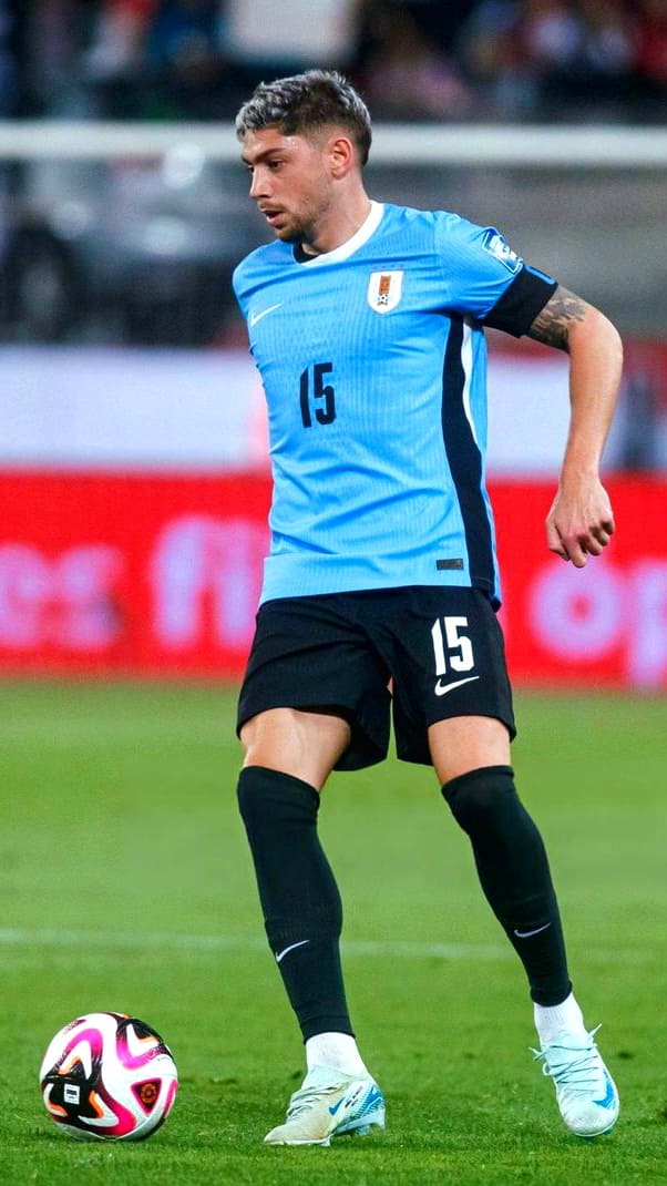 A soccer player in a light blue Uruguay jersey with the number 15 controls a ball on a green field during an IPTV Latino broadcast. The background shows blurred spectators and a red advertisement board.