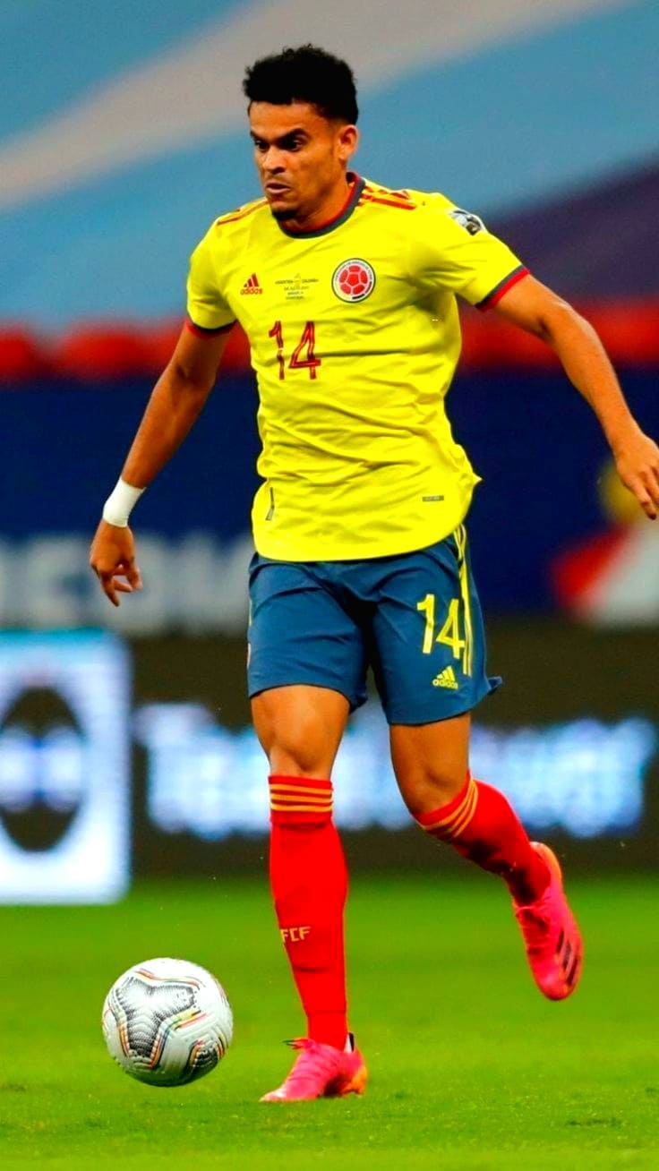 A male soccer player in a yellow Colombia jersey with number 14, blue shorts, and red socks runs on the field while dribbling a soccer ball during an IPTV Latino broadcasted match.