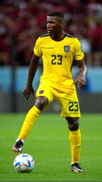 A soccer player in a yellow Ecuador national team uniform with number 23 controls the ball on a field, focused during a match. The background shows blurred spectators in a stadium, as seen live on IPTV Latino.